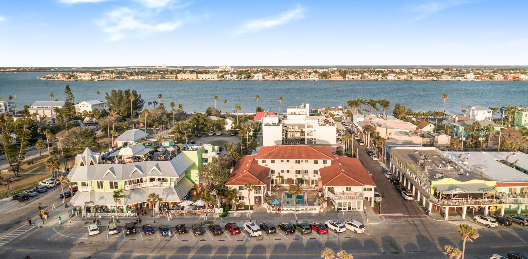 Aerial view of a waterfront neighborhood with palm trees, restaurants, parked cars, and homes along the bay.